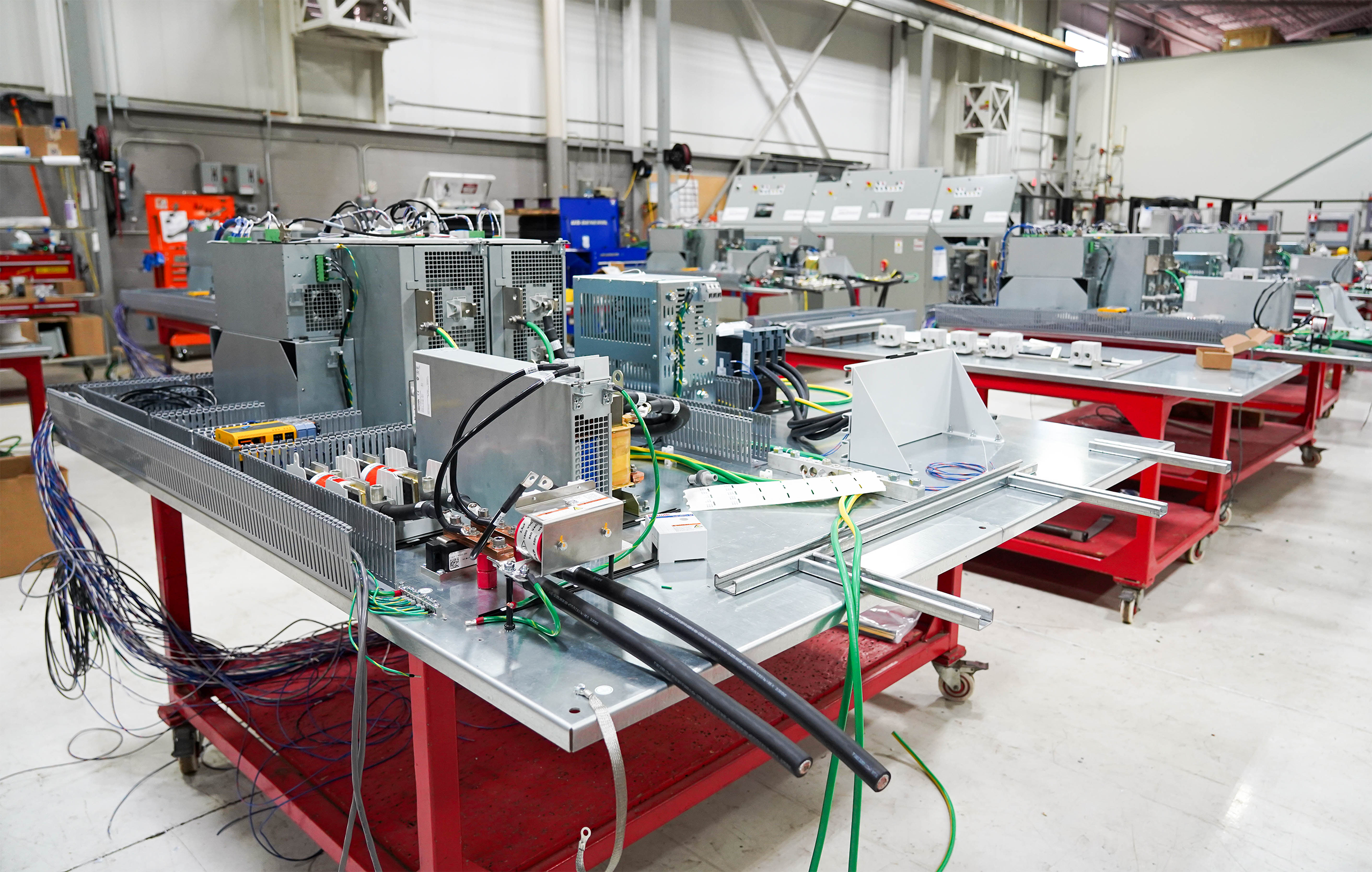Wide-angle view of an electronics assembly area with benches, power supplies, and many loose cables on red mobile carts in a manufacturing workshop