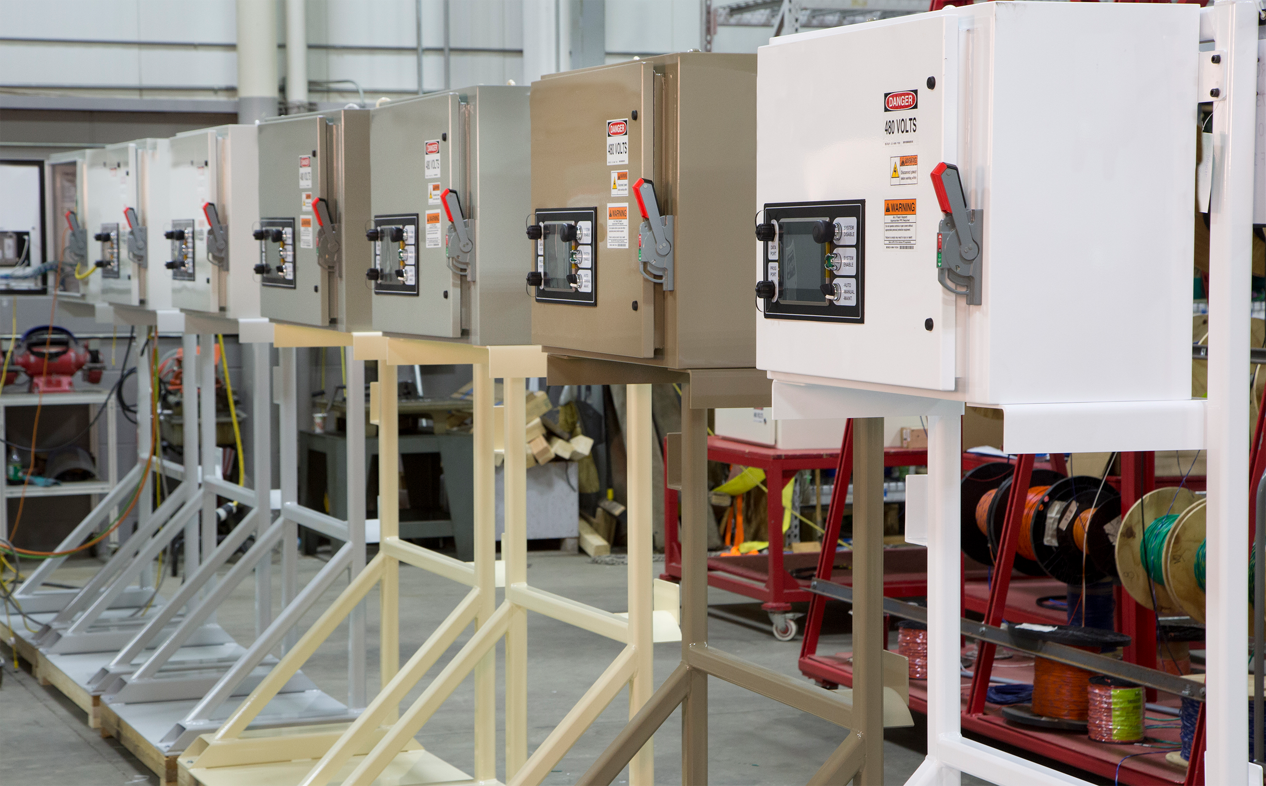Row of electrical control cabinets mounted on metal frames in a factory workshop, with warning labels and control panels visible on each unit.